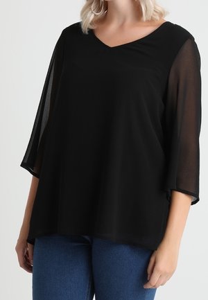 Femme portant un chemisier noir à col en V avec des manches trois-quarts transparentes et un jean bleu, debout devant un fond clair.