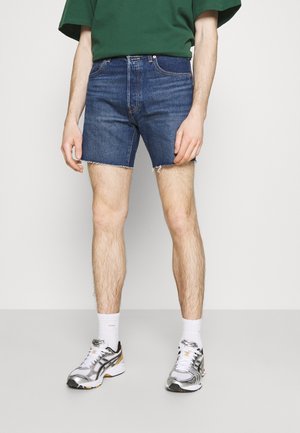 Short en jean bleu foncé avec un ourlet effiloché ; associé à des chaussures de sport grises et argentées, des chaussettes blanches et un t-shirt vert surdimensionné.