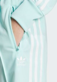 Lichtblauwe sporttrui met een ritszak, drie witte strepen op de mouw en een geborduurd logo in het wit.
