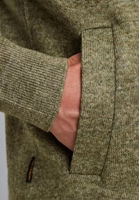 Olivengrøn strikket beklædning med en struktureret overflade; en hånd strækker sig ind i en sidelomme; har ribbet mansjettdetalje.
