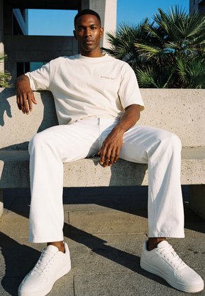 Jeune homme en T-shirt blanc et pantalon assis sur un banc en béton avec des palmiers en arrière-plan, la lumière du soleil projette des ombres.