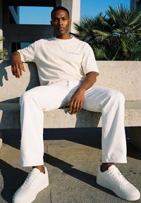 Mladi moški v belem T-shirtu in hlačah sedi na betonski klopi z rastlinami palm v ozadju, sončna svetloba meče sence.