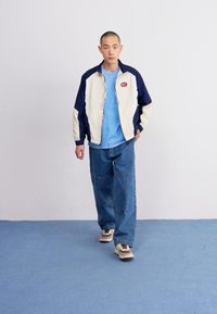 Herrfärgad nylonjacka med blockfärger i kräm och marinblå, kombinerad med en ljusblå T-shirt och lösa denimbyxor. Innehåller en rund logotyp-patch.