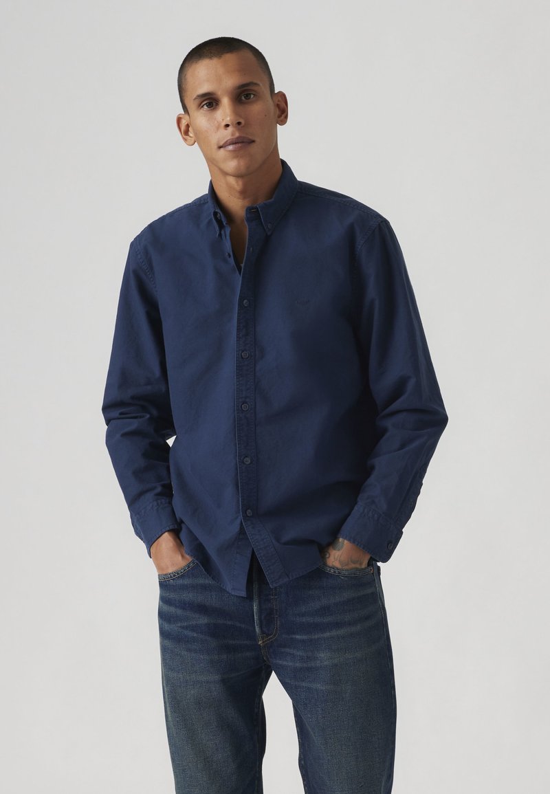 Joven con camisa azul marino de manga larga abotonada y vaqueros azules, de pie con las manos en los bolsillos frente a un fondo liso.