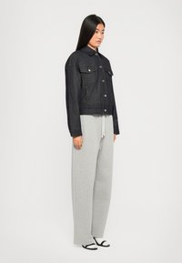 Mørk denim cropped jakke med to brystlommer, gule sømme og spids krave, parret med lysegrå sweatpants og sorte sandaler.