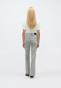 Sive teksas hlače z visokim pasom in razširjenimi hlačnicami ter črnim detajlom na obližu, kombinirane z belo majico, pogled od zadaj. Gladek tekstura.