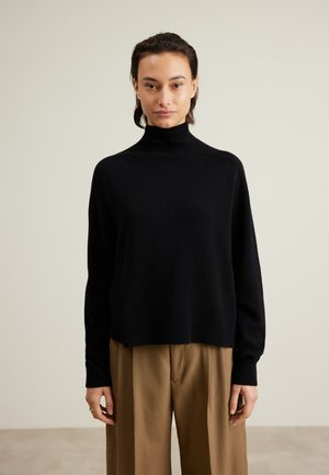 Svart turtleneck genser laget av mykt materiale, med en avslappet passform, ribbede mansjetter og nederkant. Brukt med lysebrune bukser.