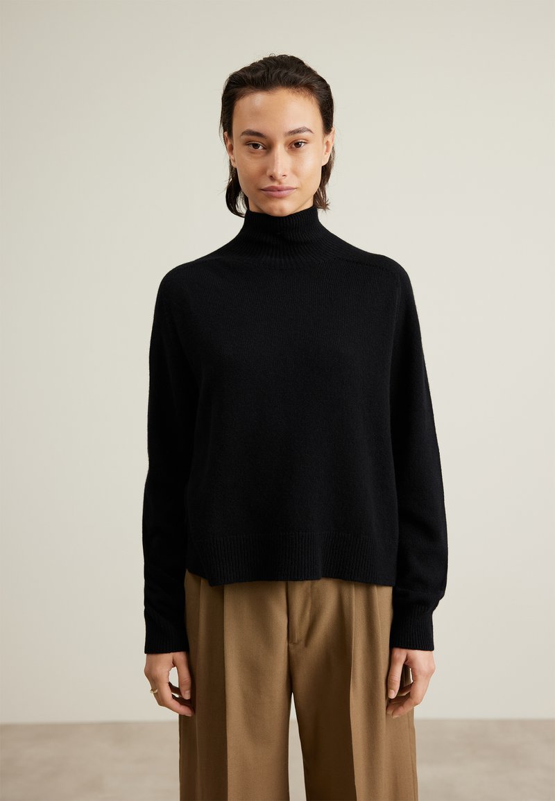 Schwarzer Rollkragenpullover aus weichem Material, mit einem entspannten Schnitt, gerippten Bündchen und Saum. Getragen mit hellbraunen Hosen.