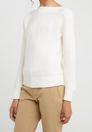 Mujer con suéter deportivo blanco de punto con puños y dobladillo acanalados, combinado con pantalones color camel, sobre un fondo claro y liso.