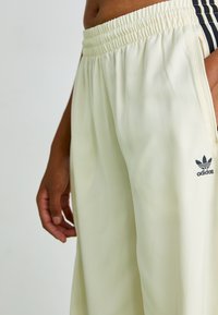 Calças de treino amarelo claro com uma cintura elástica, bolsos laterais e o logo preto da Adidas. Tecido suave com um ajuste descontraído.