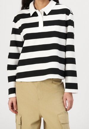 Frau trägt ein langärmliges Poloshirt mit schwarzen und weißen horizontalen Streifen und beigen Cargohosen, steht vor einem schlichten Hintergrund.