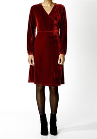 Rudé sametové zavinovací šaty s dlouhými rukávy, výstřihem do V a rozšířenou sukní. Nosit s černými kotníkovými botami přes průhledné punčochy.
