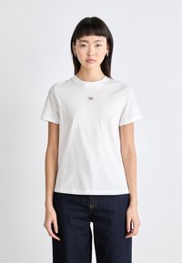 T-shirt bianca in cotone con maniche corte, scollo tondo e una piccola applicazione del logo al centro. Indossata con pantaloni in denim blu scuro.