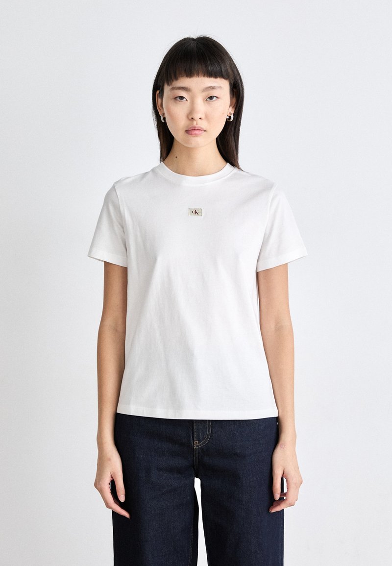 T-shirt bianca in cotone con maniche corte, scollo tondo e una piccola applicazione del logo al centro. Indossata con pantaloni in denim blu scuro.