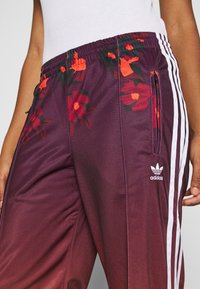 Fialové tepláky Adidas s červeným květinovým vzorem, bílými pruhy, elastickým pasem a detailem přední kapsy. Hladk�á, lehká tkanina.