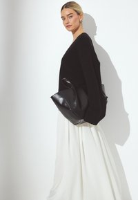 Schwarzer, übergroßer Strickpullover, kombiniert mit einem fließenden weißen Rock. Eine matte schwarze Lederhandtasche ist über die Schulter geschlungen und hat eine runde Form.