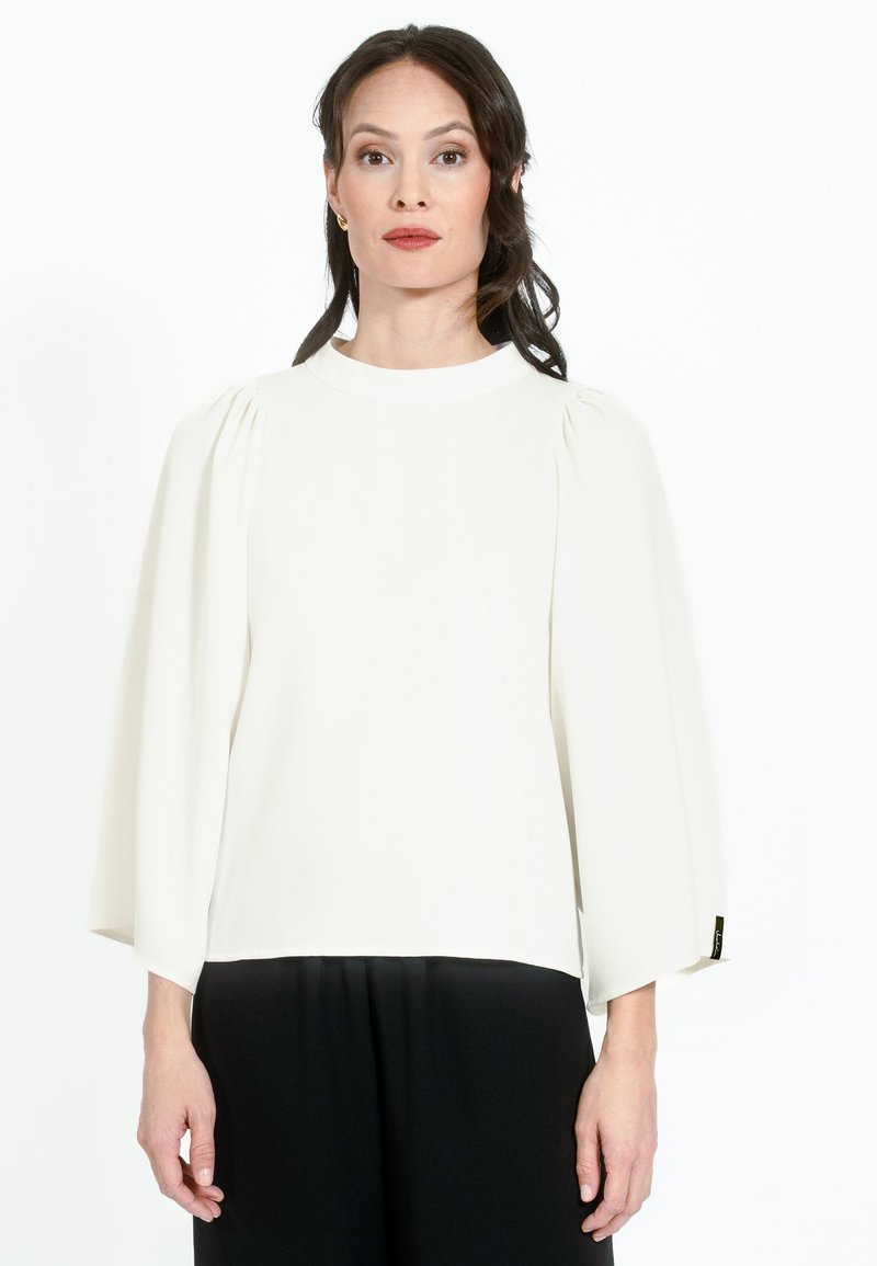 Blusa bianca a maniche lunghe con scollo rotondo e dettagli plissettati sulle spalle, abbinata a pantaloni neri, su uno sfondo semplice.