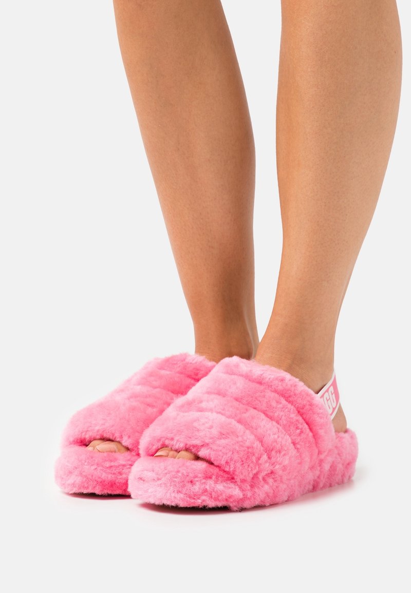 Osoba nosząca jasnoróżowe, puszyste klapki z odkrytymi palcami i paskiem z tyłu, przedstawiona z połowy łydki w dół na białym tle.