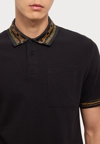 Čierna polo košela s textúrovanou látkou, so zdobným golierom v čiernej a zlatej farbe, dvoma gombíkmi a malou ľavou náprsníkou.
