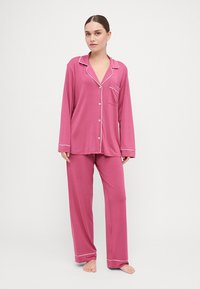 Rúžová pyžamá pozostávajúca z dlhých rukávov, košele na gombíky s vreckom a zodpovedajúcich nohavíc. Tenký biely lem dopĺňa okraje. Hladká textúra látky.