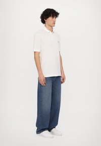 Hvid polo t-shirt med krave, parret med løse blå denimjeans og hvide sneakers. Modellen står med ansigtet mod højre foran en ensfarvet baggrund.