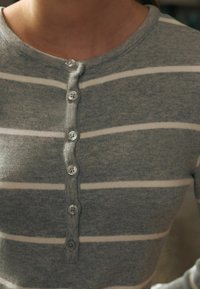 Pulover gri cu dungi albe orizontale, decolteu rotund și șase butoane pe față. Textură moale și design ajustat.