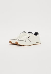 White sneakers with a smooth material, black accents, and light brown rubber sole. Features a perforated section and contrasting stitching.
