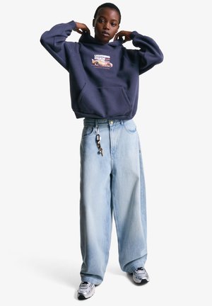 Person wearing navy hoodie with "Lightning Strikes" graphic, light blue wide-leg jeans with keys hanging, and silver sneakers, adjusting hoodie collar.