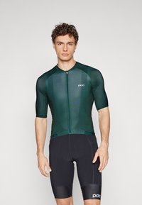 Zelený cyklistický dres s krátkymi rukávmi a predným zipsom, vyrobený z hladkého, elastického materiálu, spárovaný s čiernymi cyklistickými šortkami s logom.