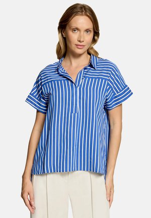 Femme à la peau claire et aux cheveux châtain clair portant une chemise bleue ample à rayures verticales blanches et un pantalon blanc sur un fond uni.