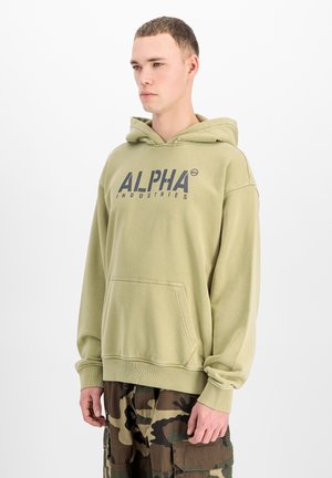 Giovane uomo che indossa una felpa con cappuccio Alpha Industries verde chiaro e pantaloni cargo mimetici, in piedi davanti a uno sfondo bianco.