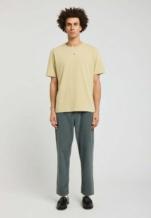 Homme aux cheveux bouclés portant un t-shirt beige, un pantalon gris, des chaussettes blanches, des chaussures noires et des bijoux en argent, debout devant un fond uni.
