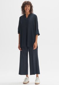 Marineblaue, locker sitzende Bluse mit V-Ausschnitt und Drei-Viertel-Ärmeln, kombiniert mit weit geschnittenen dunkelgrauen Hosen und weißen Sneakers.