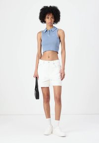 Lys blå kortermet polo med ribbet tekstur, kombinert med hvite denimshorts med frynset kant. Modellen holder en svart håndveske.