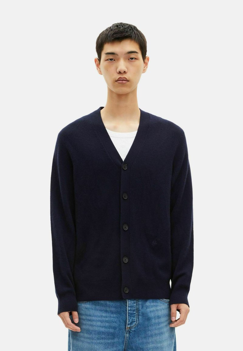 Navy blue cardigan made of soft fabric, featuring a V-neck, five buttons, and ribbed cuffs. Worn over a white t-shirt and denim jeans.