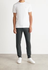 Mann in einem schlichten weißen T-Shirt mit kleinem roten II-Logo, dunklen schmalen Hosen und weißen Sneakern, der auf einem glatten Boden vor einer weißen Wand steht.