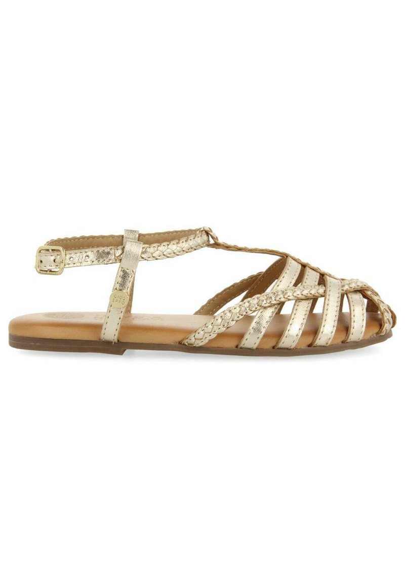Sandalias metálicas doradas con tiras trenzadas, punta abierta y suela plana. Banda lateral en el tobillo con detalle de hebilla ajustable.