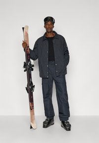 Echipamentul de schi include o jachetă în carouri închise, o cămașă neagră, pantaloni închiși și cizme de schi negre. Accesorii: ochelari de schi și o pereche de schiuri cu legături.