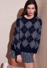 Navy and gray argyle sweater with silver detailing, round neckline, and ribbed cuffs. Paired with a pleated gray skirt.