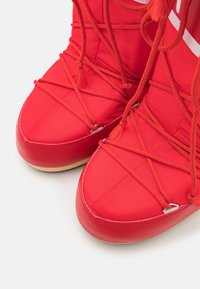 Bright red winter boots with thick fabric, rope-style laces crisscrossing the toe area, and beige soles on a white background.