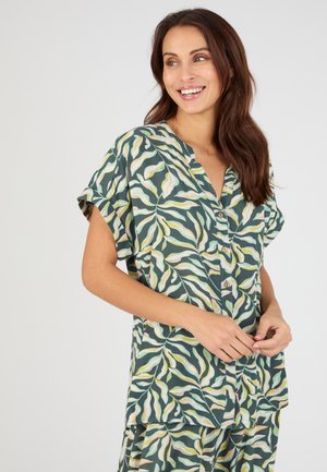 Mujer sonriente con cabello largo y castaño lleva una camisa verde abotonada y pantalones a juego con un patrón de hojas en amarillo, blanco y rosa.