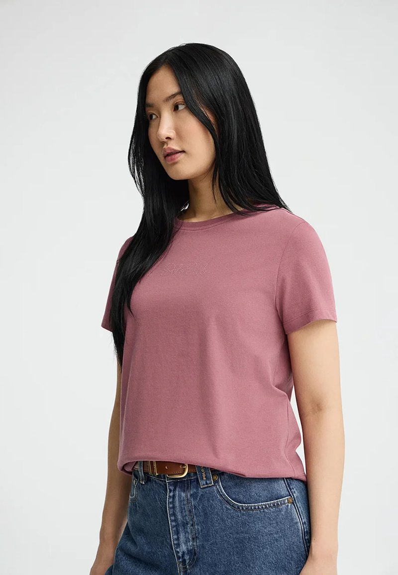 Mujer joven con cabello largo y negro, vestida con una camiseta de manga corta color malva metida dentro de unos jeans azules de cintura alta y un cinturón marrón.