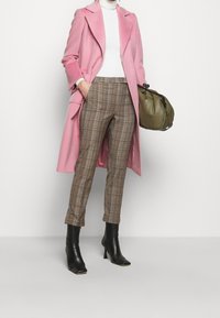 Manteau en laine rose, col roulé blanc, pantalon à carreaux marron, et bottines à talons noires, avec un sac à main vert à la main.