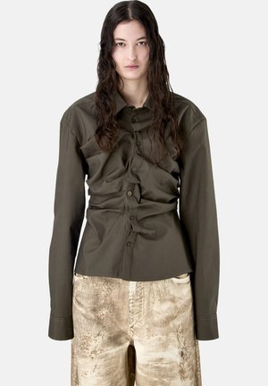 Jonge persoon met lang, nat haar draagt een olijfgroen overhemd met knopen en beige broek met slijtage, staand tegen een effen achtergrond.