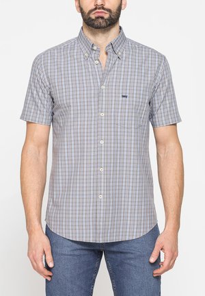 Uomo che indossa una camicia a maniche corte a quadri nelle tonalità di grigio e blu con jeans blu, in piedi davanti a uno sfondo bianco semplice.