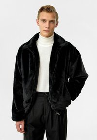 Zwarte faux fur zip-up jacket met een zachte textuur, grote kraag en drop shoulders, gecombineerd met een lichte trui en op maat gemaakte broek.