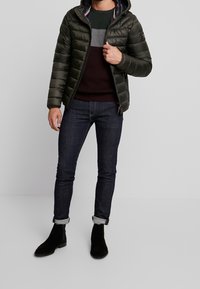 Daunenjacke in Dunkelgrün, gesteppter Schnitt mit Kapuze. Kombiniert mit dunklen Jeans und schwarzen Wildleder-Stiefeletten. Akzentuiert durch einen mehrfarbigen Pullover.