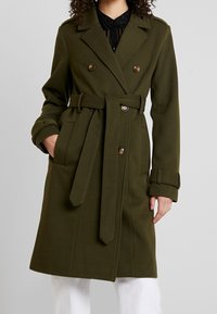 Olijfgroene, dubbelgeknoopte trenchcoat met een ceintuur in de taille, gestructureerde stof en knoopdetails aan de voorkant en op de mouwen.