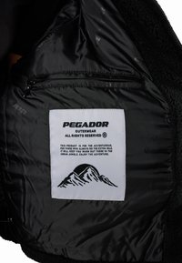 Un capo di abbigliamento esterno nero con un morbido rivestimento in felpa e una tasca interna. Un'etichetta bianca mostra la scritta "PEGADOR OUTERWEAR" con una grafica di una montagna.