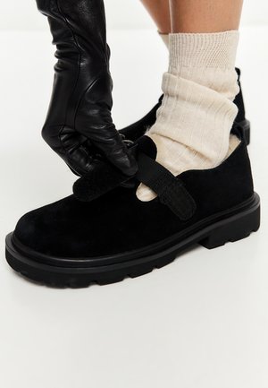 Hånd i sort læderhandske, der spænder velcrobånd på sort sko båret med cremefarvede ribstrikkede sokker.